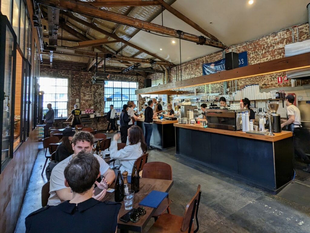 Large, open cafe space, with distressed brick walls and high ceilings with exposed wooden beams. Long counter runs down one side, with several staff members behind. Line of people waiting for coffee at one end, with a few tables nearby.