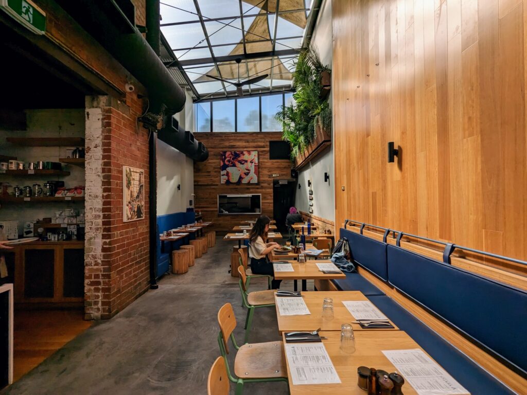 Interior view of a bright, industrial cafe in what looks like a converted warehouse with some original brick walls visible. Several tables and chairs (mostly empty) run along a wooden wall. Coffee equipment visible on shelves to the left. High windows and skylights above.