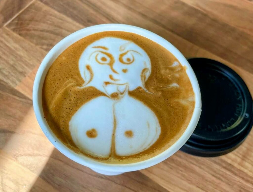 Overhead view of takeaway coffee cup with latte art depicting a woman with large breasts, sitting on a wooden table. Black lid for takeaway cup is alongside.