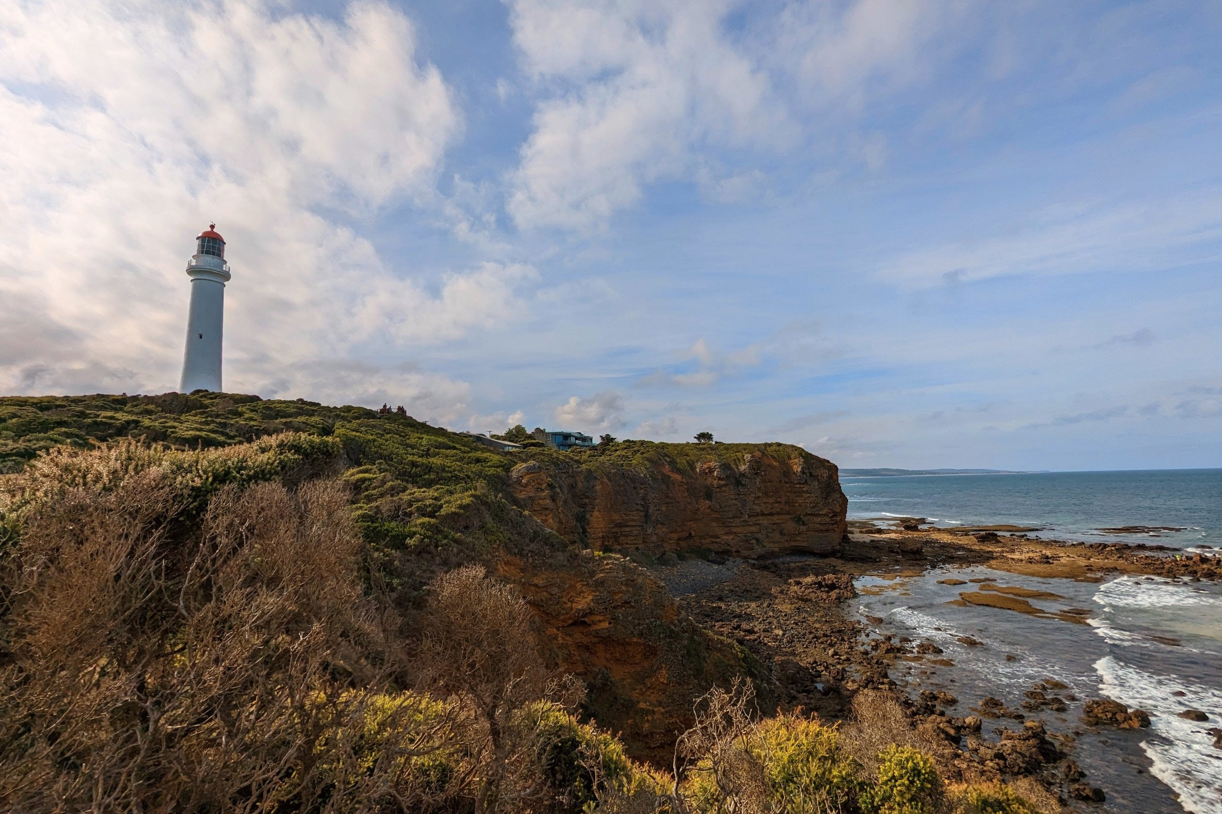 Lighthouse on weathered cliff face, overlooking rocky ocean bay below. Trees in the foreground, blue sky with white clouds above