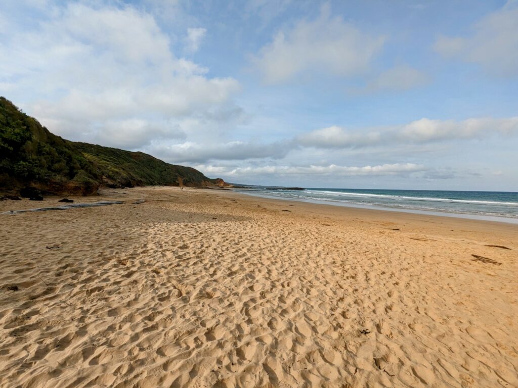 Wide stretch of golden beach with sandstone cliffs behind. Blue sky and clouds above.