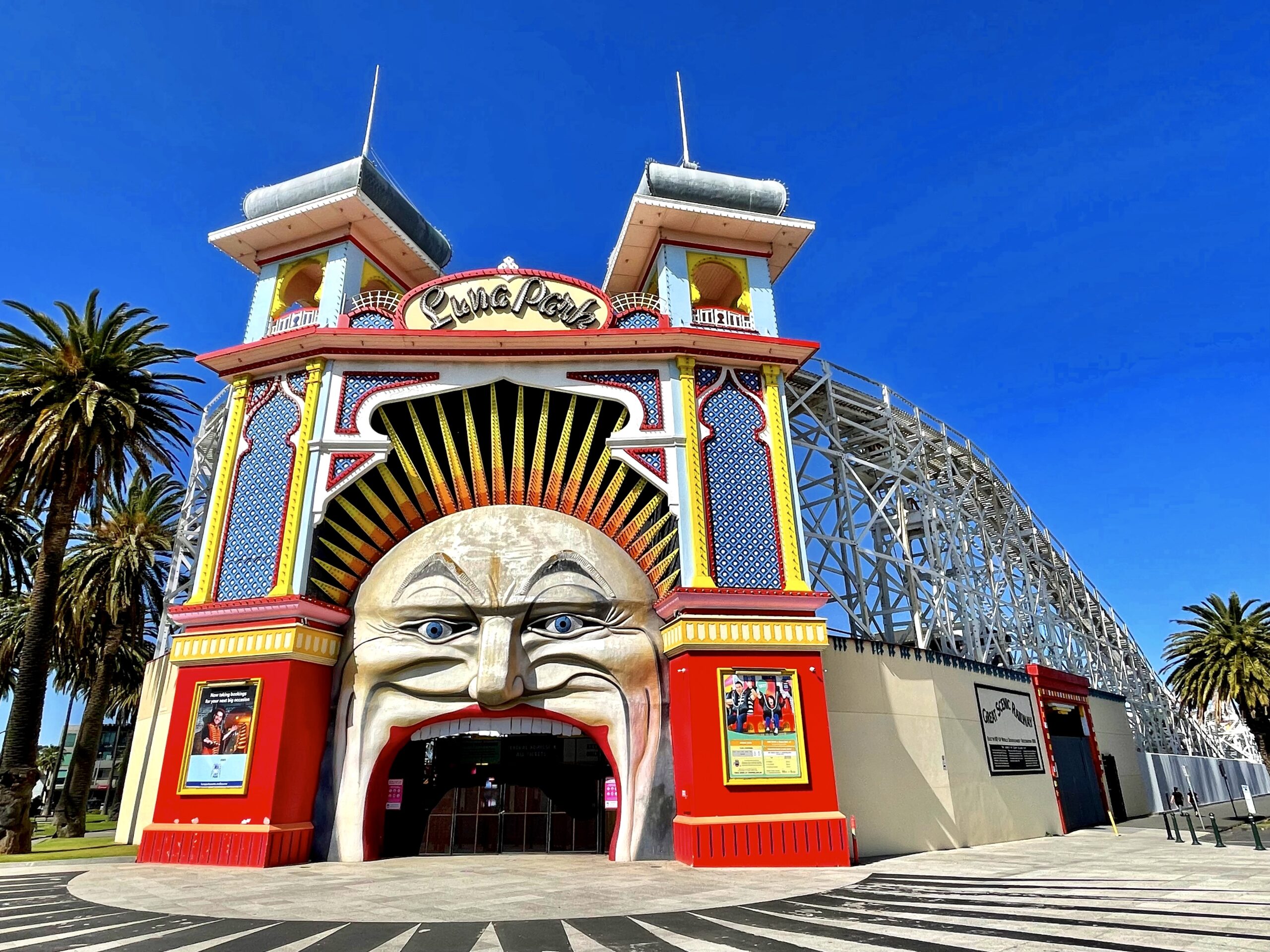 The retro, Art-Deco-themed entrance to St Kilda's Luna Park, featuring an enormous face of a clown with a door providing the entrance into the amusement park. The tracks of a wooden roller coaster form the perimeter of the park.