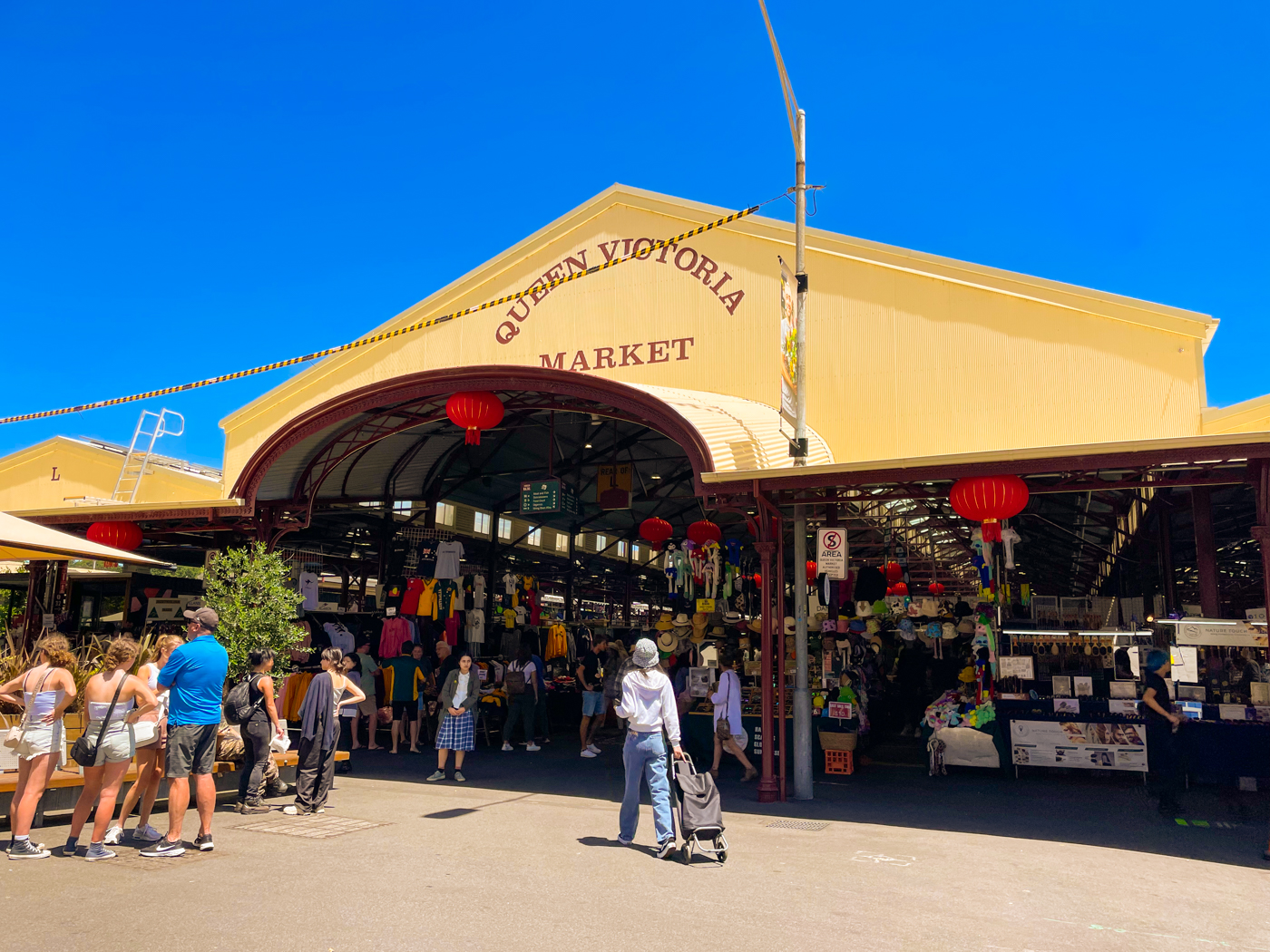 A quiet entrance to the Queen Victoria Market on a sunny, cloudless day. The faded yellow facade has the name of the market on it, while the shaded interior shows souvenir stalls for tourists to browse.