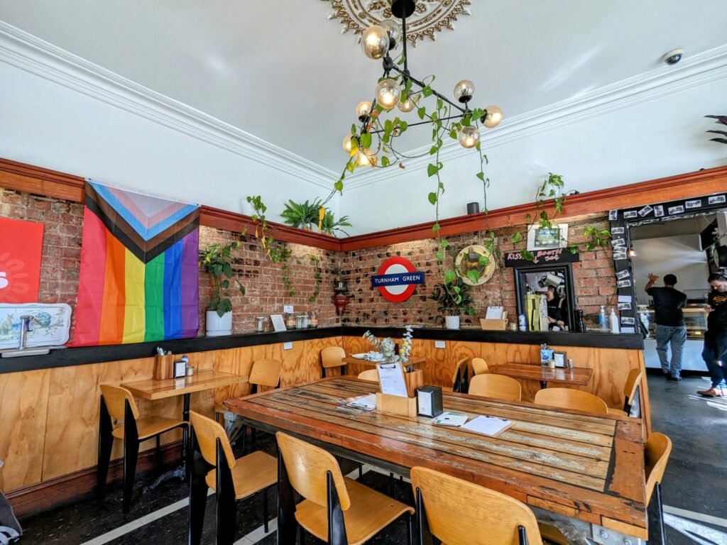 Interior of a cafe ,with wooden tables and chairs. Walls are wood on the bottom half and brick on the top half. Rainbow flag on one wall, Turnham Green London Underground sign on another. Small window cut out of one wall with staff visible making coffee on the other side.