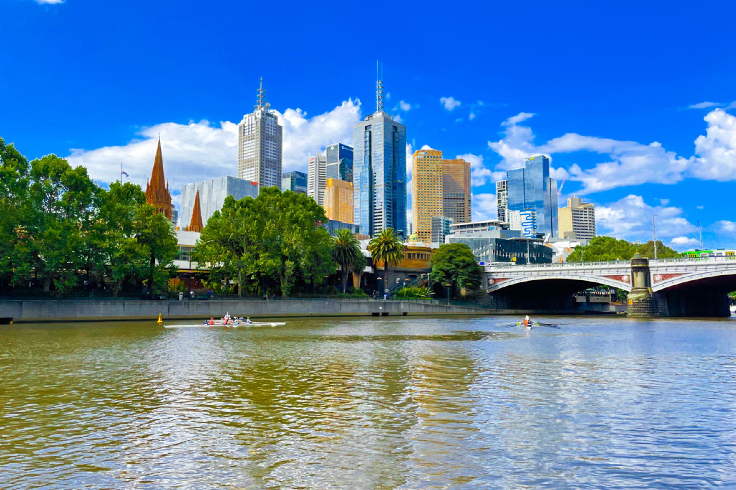 In the distance, the skyscrapers of Melbourne's CBD reflect the sunlight and blue sky on a sunny day. In front of the buildings, the murky Yarra River passes beneath a bridge.