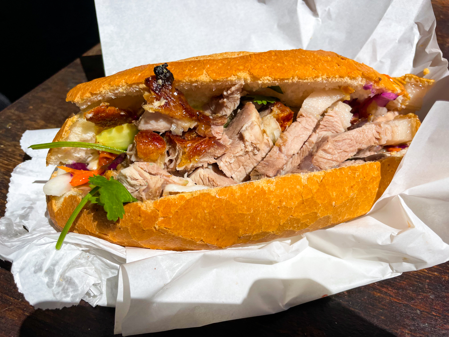 A crispy pork banh mi from T&L Bakery in Cremorne. The paper wrapper is splayed out across a wooden table with the crunchy sandwich placed in the center. Crispy, fatty pork chunks are seen spilling out of the banh mi, with hints of coriander, carrot, and cucumber poking out of the sides.