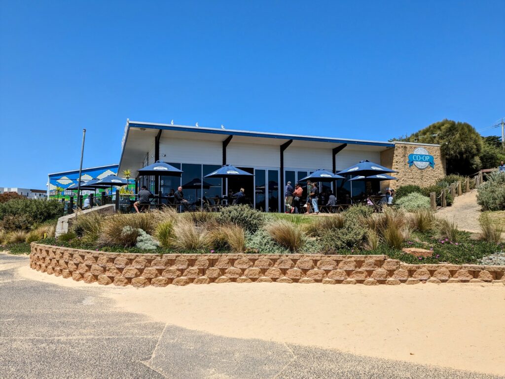 Exterior view of the San Remo Fisherman's Co-Op building in San Remo, with outdoor tables shaded by umbrellas visible. A small garden with native plants in front, and a mix of concrete and sandy path.