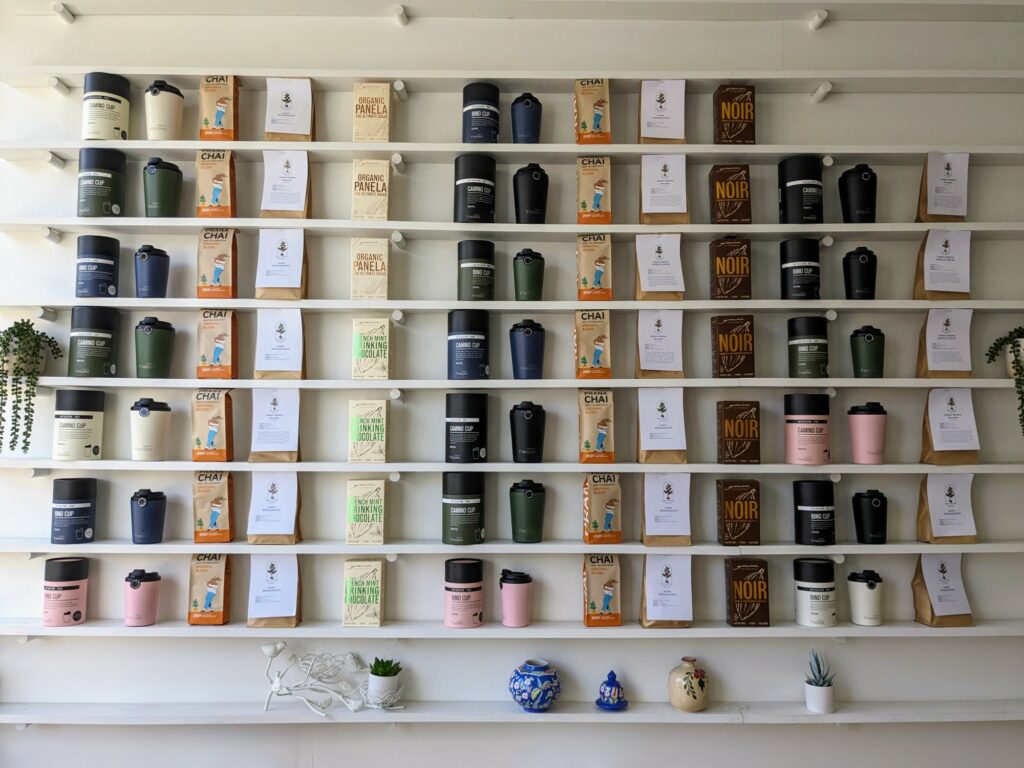 Several shelves mounted on a white wall with bags of coffee, chair, and drinking chocolate, plus takeaway cups and storage canisters, artfully arranged on them