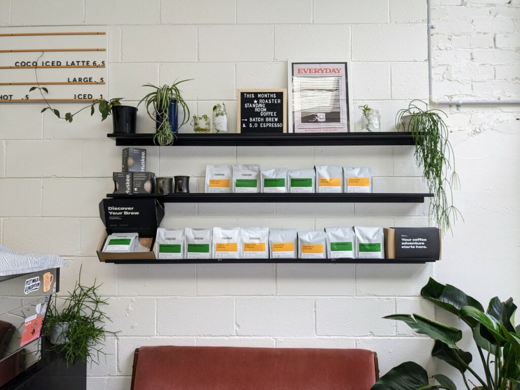 Shelves with bags of coffee beans and coffee equipment, mounted on a white brick wall. Plants visible nearby, along with a partial coffee menu and espresso machine.