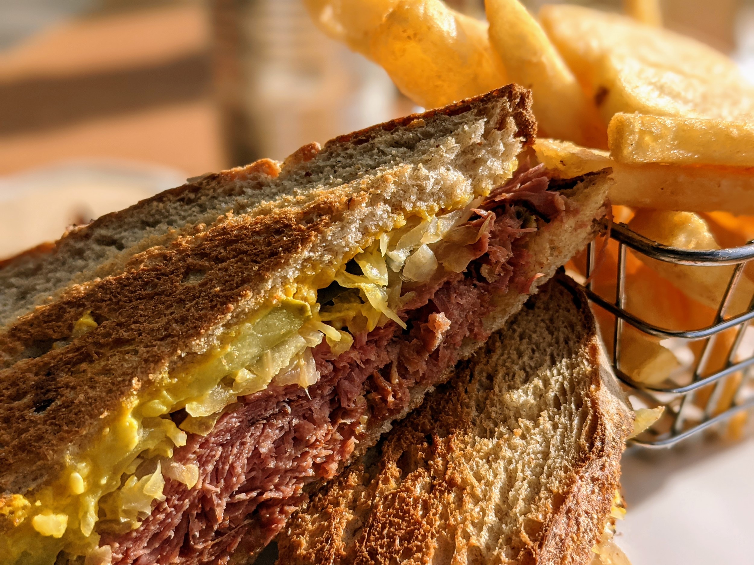 Closeup of part of a reuben sandwich with toasted brown bread, with a basket of fries in the background