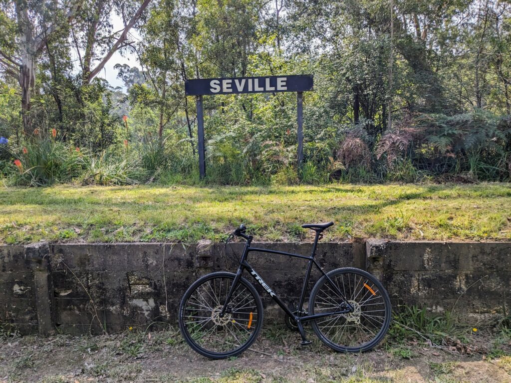 Bicycle leaning up against the concrete side of an old railway platform, now covered in grass. A raised sign reading "Seville" is at the rear of the platform.