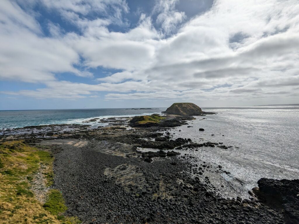 View from the shore over a rocky beach towards outcroppings of land that rise up from the ocean just offshore. Patches of blue sky in between thick white cloud.