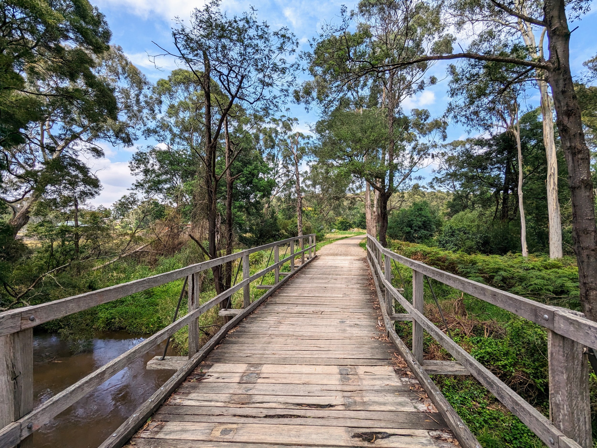 Wooden bridge over a small brown river with bushes and trees on both sides.