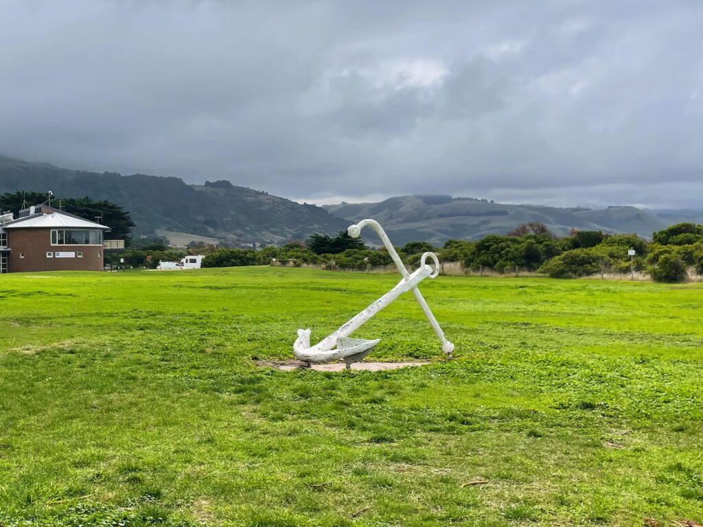 White-painted ship's anchor sitting in a grassy park, with hills in the background.
