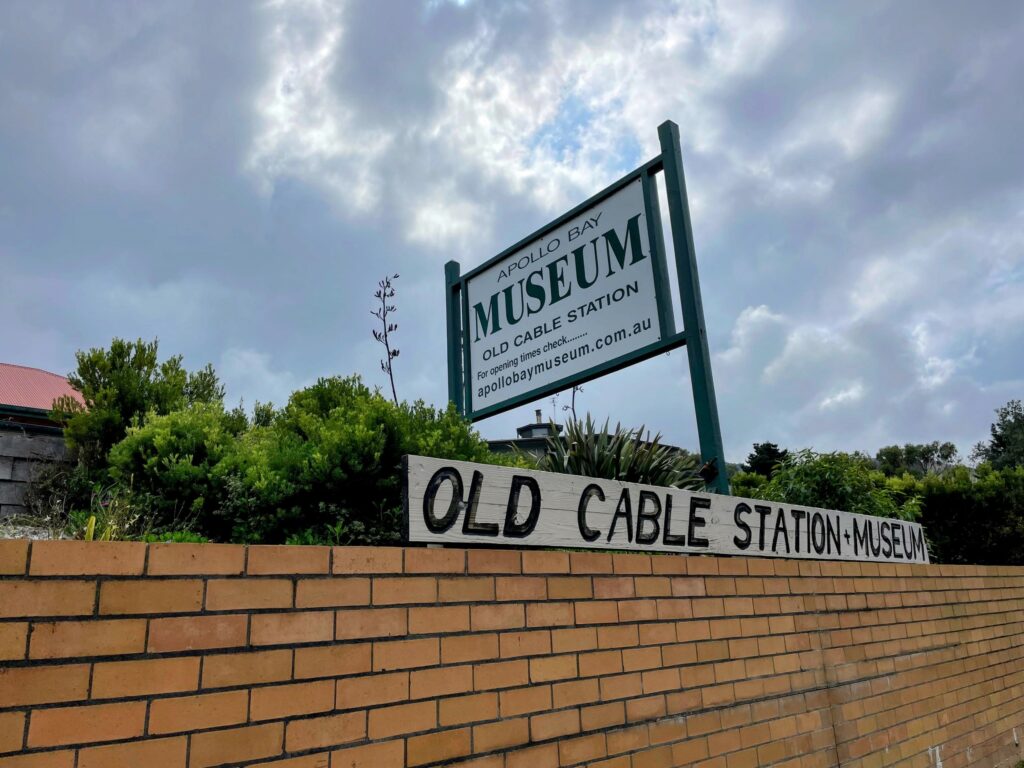 Exterior of the Apollo Bay museum, with a brick wall above which sits a hand-painted sign reading "Old cable station+museum", behind which sits a printed sign saying "Apollo Bay Museum"