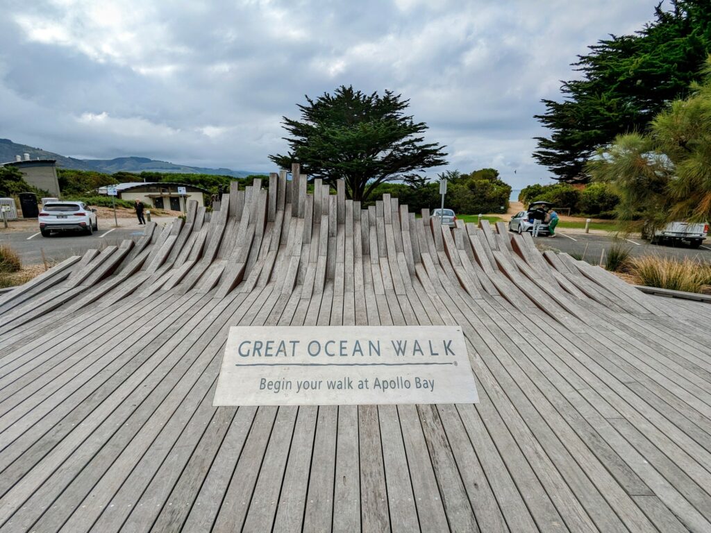 Large wooden decking sculpture curved up at the ends to resemble a wave, with a sign inlaid in it that says "Great Ocean Walk: Begin your walk at Apollo Bay". Trees and a small car-park behind