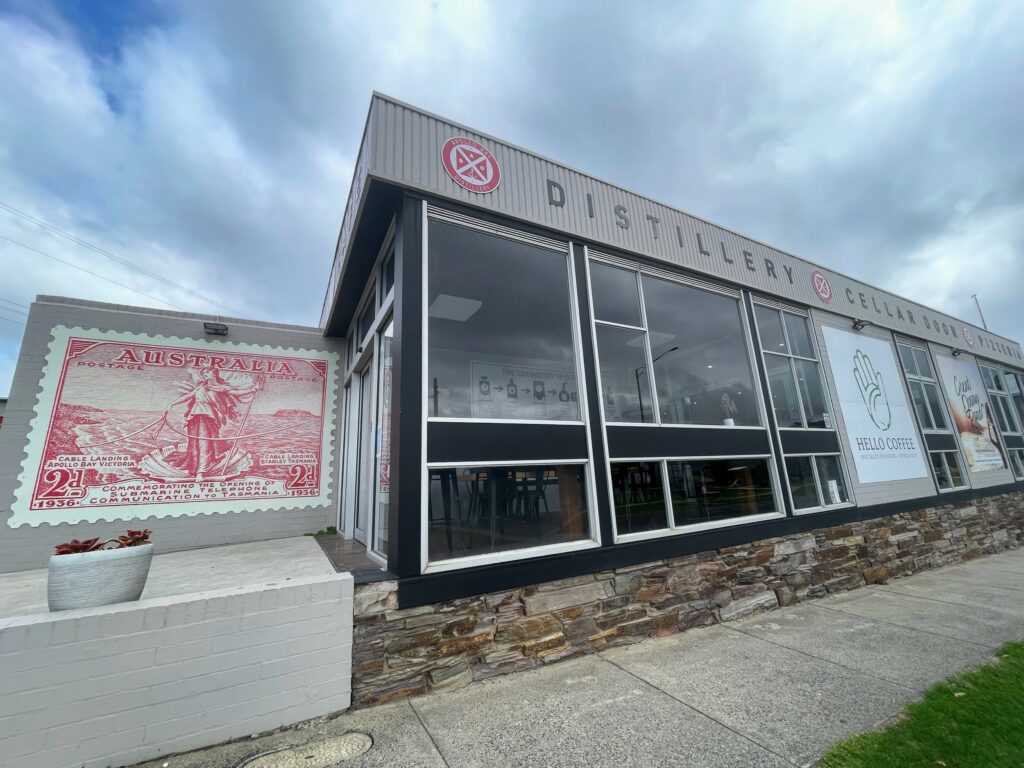 Outside of a glass-fronted building with signage saying "Distillery - Cellar Door - Pizzeria" above the windows. A large replica of a 1936 two-penny Australian stamp is printed on the wall.