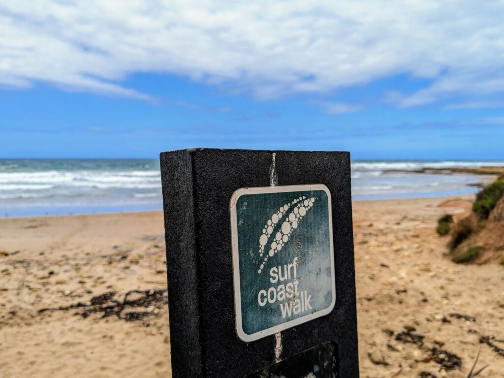 Wooden signpost with a stylised leaf symbol and the words "Surf Coast Walk" printed underneath. A beach and ocean are visible behind.