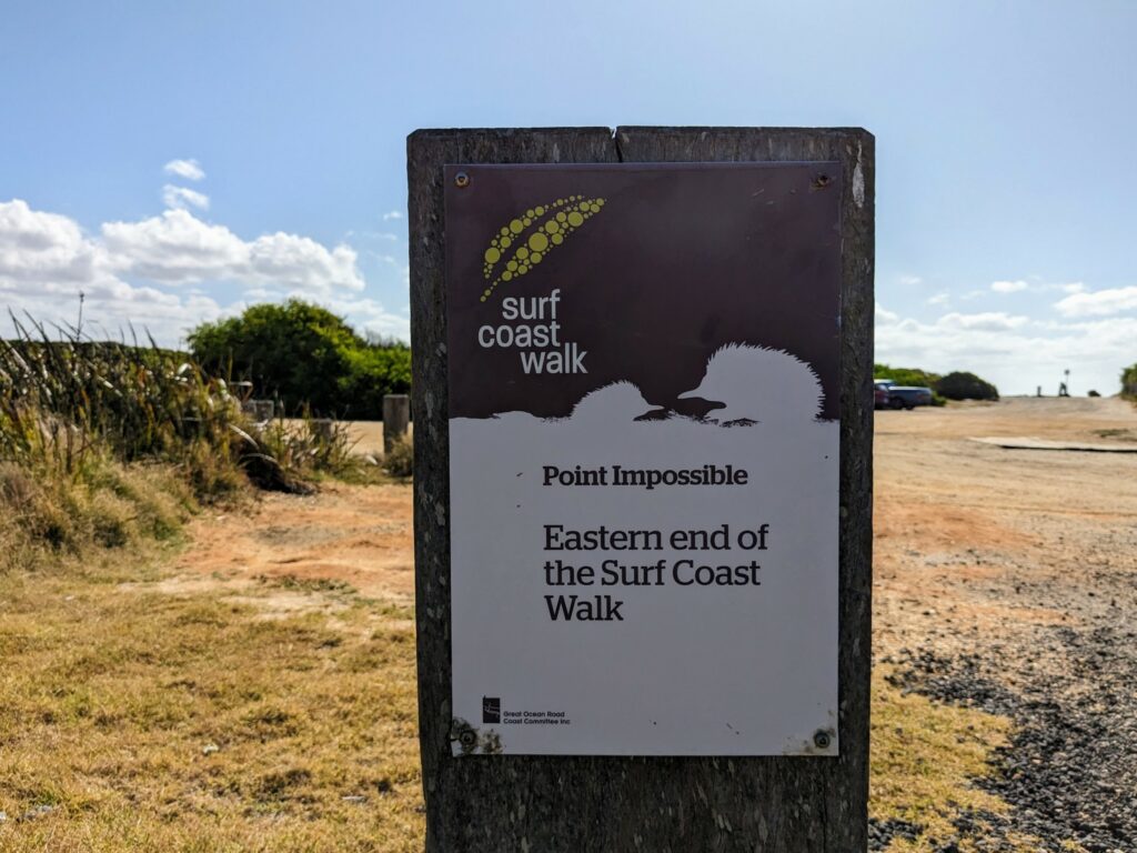 Wooden signpost with printed sign attached saying "Surf Coast Walk. Point Impossible. Eastern end of the Surf Coast Walk". A dirt carpark is visible behind.
